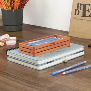 May include: A wooden pencil holder with the word "WOODSAGA" engraved on the front. The holder is filled with blue and green colored pencils and is sitting on top of two notebooks.