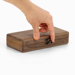 May include: A rectangular wooden box with a dark brown finish and a metal clasp. The box is closed, and a hand is reaching towards the clasp. The wood grain is visible on the surface of the box.