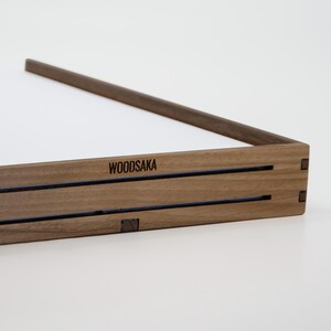 May include: A wooden tray with a dark brown finish and the word "WOODSAKA" engraved on the front. The tray has a simple design with two parallel lines running across the front.
