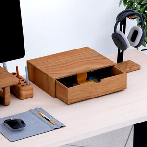 May include: A wooden desk organiser with an open drawer, displaying a warm, natural wood grain. The drawer reveals office supplies. A mouse, pen, and a small wooden pen holder are also on the desk. Headphones hang on a black stand.