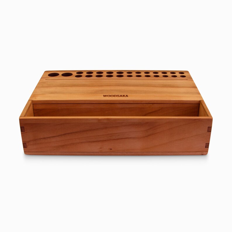 May include: A wooden box with a lid that opens to reveal a compartment. The box has a row of holes on the top and the word "WOODSAKA" is engraved on the front.