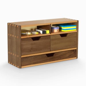 May include: A wooden desk organiser with three drawers and a top shelf with dividers. The organiser is made of walnut wood and has a natural finish. The top shelf is filled with office supplies, including pens, pencils, and tape.