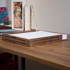 May include: A wooden desk organizer with a black stripe design. The organizer has the text "WOODSARA" engraved on the front. The organizer is sitting on a wooden desk.