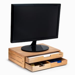 May include: A wooden monitor stand with two drawers. The stand is made of light brown wood and has a natural finish. The drawers are labelled "WOODENAKA".