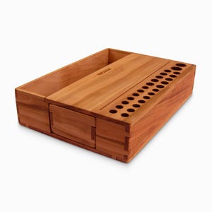 May include: A wooden organizer with a compartment for storing items and a row of holes for holding pens or pencils. The organizer is made of cherry wood and has a natural finish.