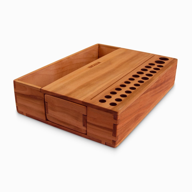 May include: A wooden organizer with a compartment for storing items and a row of holes for holding pens or pencils. The organizer is made of cherry wood and has a natural finish.