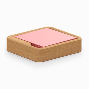 May include: A square wooden desk organizer with rounded corners and a light brown wood grain finish. The center of the organizer features a stack of square, solid light pink paper notes that sit flush within the wooden frame.