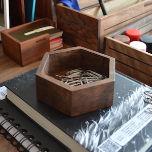 May include: A hexagonal wooden bowl with a dark brown finish holds a pile of silver paper clips. The bowl is sitting on top of a black book with a white design.