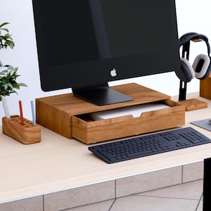 May include: A wooden desk setup with a black computer monitor, a wooden monitor stand with a drawer, a black keyboard, and a wooden pen holder. The desk also includes headphones and a small plant. The monitor stand is approximately 30 cm wide.