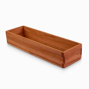 May include: A long, rectangular wooden box with a natural cherry finish. The box is made of solid wood and has a smooth, polished surface.