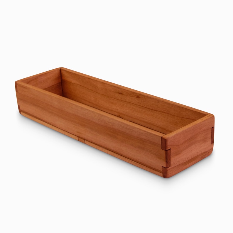 May include: A long, rectangular wooden box with a natural cherry finish. The box is made of solid wood and has a smooth, polished surface.