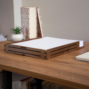 May include: A wooden desk organizer with the text "WOODSAKA" on the front. The organizer is made of dark brown wood and has a rectangular shape. It is sitting on a wooden desk with a stack of books and a potted plant in the background.