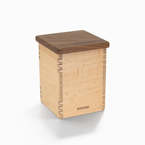 May include: A square wooden box with a walnut lid and maple sides. The box has a simple design and is labeled "WOODSAKA" on the front.
