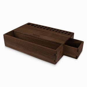 May include: A wooden organizer with a drawer and a compartment with multiple holes for storing pens and pencils. The organizer is made of dark walnut wood and has the word "Remarkable" engraved on the front.