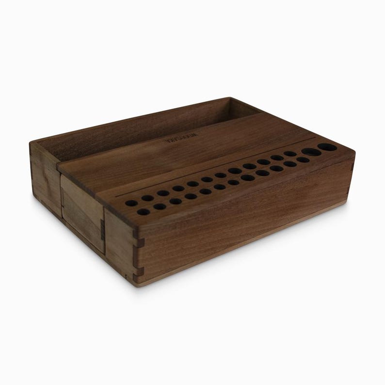 May include: A wooden organizer with a compartment for a phone and a row of holes for storing pens or pencils. The organizer is made of dark wood and has a natural finish.