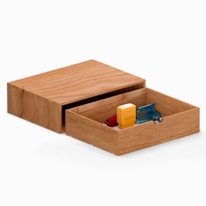 Puede incluir: Una caja de madera con un cajón corredero, parcialmente abierto para revelar pequeños útiles de oficina. La caja es de madera clara con veta visible. Dentro del cajón hay una grapadora roja, un sello amarillo y una perforadora azul.