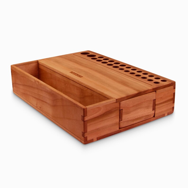 May include: A wooden organizer with a compartment for storing items and a section with multiple holes for holding pens or pencils. The organizer is made of light brown wood and has the word "WOODSHED" engraved on the top.
