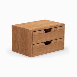 Peut inclure: Un coffret de rangement en bois à deux tiroirs avec une finition en bois naturel. La boîte présente des assemblages à queue d'aronde et des poignées de tiroir encastrées. Les tiroirs sont vides, révélant un aperçu des intérieurs bleus et noirs.