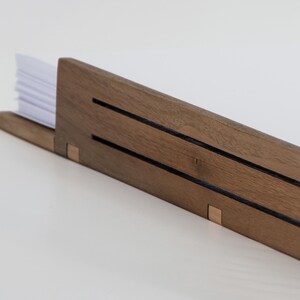 May include: A wooden desk organizer with two slots for holding papers. The organizer is made of dark brown wood with a natural grain pattern. The organizer has a simple, minimalist design.