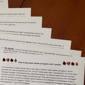 May include: A stack of white paper sheets with printed text, likely instructions for a card game. The text includes details on how to play Super Samba with two people, card trays, and card caddies. The papers are on a wooden surface.