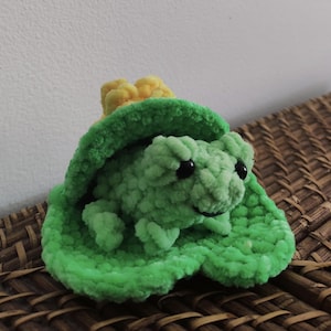 May include: A handmade, plush frog in a bright green hue, nestled on a matching lily pad. The frog has black eyes and is accented with a yellow flower. The soft, textured yarn gives the toy a cuddly appearance.