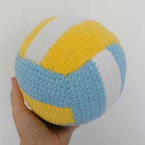 Könnte beinhalten: Ein handgehäkelter Volleyball in Gelb, Weiß und Hellblau. Der Ball wird in einer Hand vor einem weißen Hintergrund gehalten. Das Design des Balls ahmt einen echten Volleyball nach.