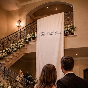 May include: A couple stands in an elegant hall, looking up at a banner that reads "The Mc Cues." The scene features a grand staircase adorned with white flowers and candles, creating a romantic ambiance. The woman wears a sparkling gown, and the man is in a black suit.