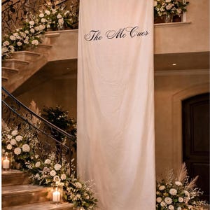 May include: Elegant wedding decor featuring a staircase adorned with white flowers and greenery. A large white banner with the text "The Mc Cues" hangs in the center. Lit candles add a warm glow.