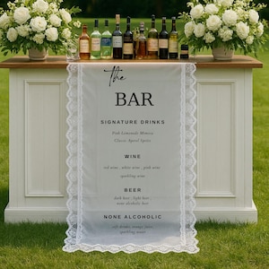 Può includere: Un bar bianco con un piano d'appoggio in legno, decorato con composizioni floreali bianche e dettagli di cristallo sospesi. Il bar presenta un'insegna con la scritta "The Bar" e elenca drink speciali, vini, birre e opzioni analcoliche. Sono esposte diverse bottiglie.
