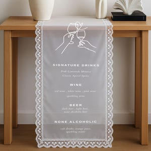 Pode incluir: Um caminho de mesa branco transparente com uma delicada borda de renda, apresentando um menu de bebidas impresso. O menu inclui bebidas exclusivas, vinho, cerveja e opções não alcoólicas. Um vaso com flores brancas e objetos decorativos estão na mesa.