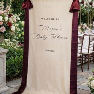 May include: A cream-colored banner with burgundy ribbons and text that reads "WELCOME TO Megan's Baby Shower" and the date "20 07 2025". The banner is displayed outdoors, with floral arrangements and a table setting in the background.