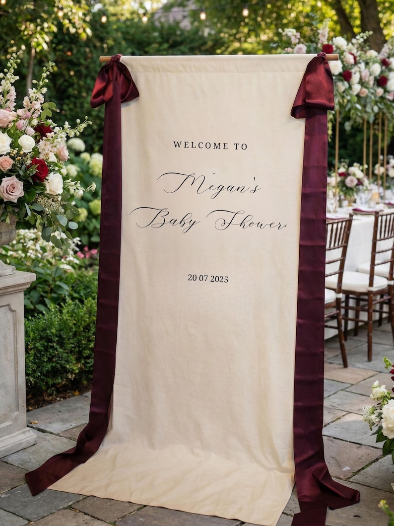 May include: A cream-colored banner with burgundy ribbons and text that reads "WELCOME TO Megan's Baby Shower" and the date "20 07 2025". The banner is displayed outdoors, with floral arrangements and a table setting in the background.