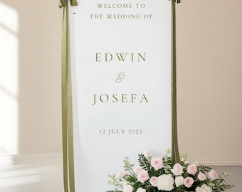 May include: A white wedding welcome sign with green text that reads "Welcome to the Wedding of Edwin & Josefa 12 July 2025." The sign is decorated with olive green ribbons and a floral arrangement of pink and white roses.