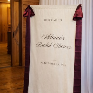 May include: A cream-colored banner with burgundy ribbons and accents, announcing "Welcome to Melanie's Bridal Shower" with the date November 15, 203. The banner is hanging in front of a white curtain.