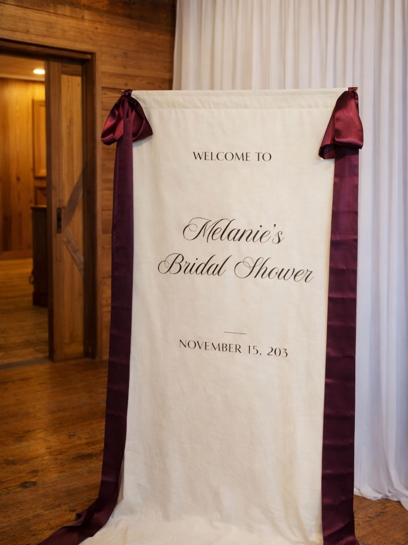 May include: A cream-colored banner with burgundy ribbons and accents, announcing "Welcome to Melanie's Bridal Shower" with the date November 15, 203. The banner is hanging in front of a white curtain.