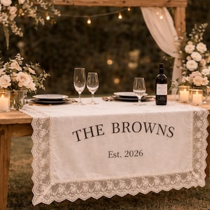 Puede incluir: Una mesa de madera rústica preparada para una boda, adornada con un camino de mesa blanco con bordes de encaje y el texto "THE BROWNS Est. 2026". La mesa está puesta con platos, copas de vino y una botella de vino. Arreglos florales y velas crean un ambiente romántico.