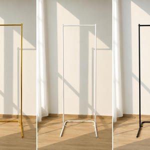 May include: Three metal clothing racks in gold, white, and black. Each rack has a rectangular frame with a horizontal bar at the top and a base for stability. The racks are displayed against a white wall with natural light.