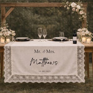 May include: A wedding table setting with a wooden frame and floral decorations. A white lace runner displays "Mr. & Mrs. Matthews" and the date "11.08.2025". The table is set with plates, champagne flutes, and candles for the reception.