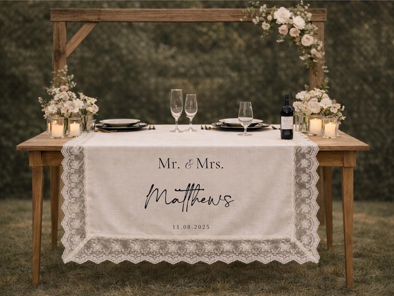 May include: A wedding table setting with a wooden frame and floral decorations. A white lace runner displays "Mr. & Mrs. Matthews" and the date "11.08.2025". The table is set with plates, champagne flutes, and candles for the reception.