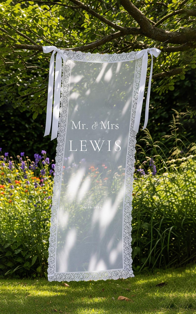 K&ouml;nnte beinhalten: Ein durchsichtiges wei&szlig;es Hochzeitsbanner mit zarten Spitzenbesatz und wei&szlig;en B&auml;ndern, das an einem Ast h&auml;ngt. Das Banner zeigt die Namen "Mr. & Mrs. LEWIS" und das Datum "06.26.2025".