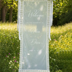 K&ouml;nnte beinhalten: Ein durchsichtiges wei&szlig;es Hochzeits-Willkommensschild mit zarten Spitzenborten und wei&szlig;en B&auml;ndern. Auf dem Schild stehen die Worte "Welcome" und "Olivia + Davis" mit dem Datum "May 16, 2024". Das Schild h&auml;ngt an einem Ast in einem Feld.