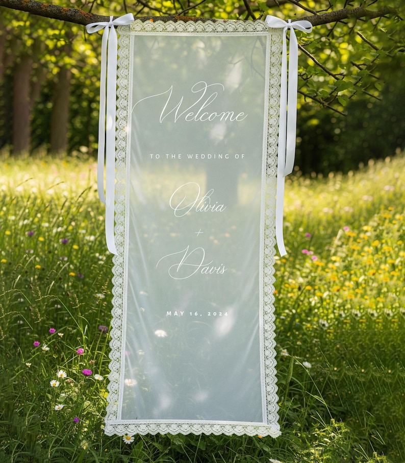 K&ouml;nnte beinhalten: Ein durchsichtiges wei&szlig;es Hochzeits-Willkommensschild mit zarten Spitzenborten und wei&szlig;en B&auml;ndern. Auf dem Schild stehen die Worte "Welcome" und "Olivia + Davis" mit dem Datum "May 16, 2024". Das Schild h&auml;ngt an einem Ast in einem Feld.
