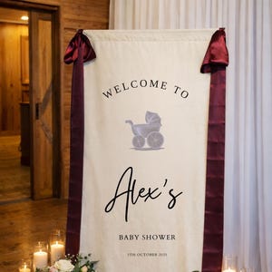 May include: A cream-colored baby shower banner with the text "WELCOME TO Alex's BABY SHOWER 5TH OCTOBER 2025" and a stroller graphic. The banner is adorned with burgundy ribbons and bows. Floral arrangements and candles flank the banner.