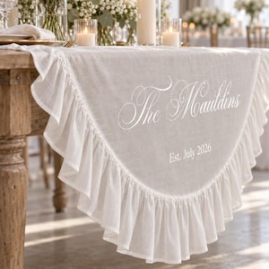 May include: A white semi-circular table runner featuring a ruffled edge and white embroidered text that reads The Mauldins and Est. July 2026. The fabric has a visible weave texture and drapes over the edge of a wooden table.