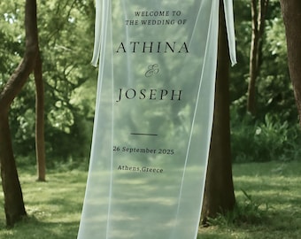 May include: A sheer white wedding banner suspended between trees. The banner reads "WELCOME TO THE WEDDING OF ATHINA & JOSEPH" with the date "26 September 2025" and location "Athens, Greece" in black text.