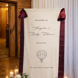 May include: A cream-colored baby shower welcome banner with burgundy ribbon accents. The banner reads "Welcome to Angelee's Baby Shower" with the date 04.26.2025 and a hot air balloon graphic. Floral arrangements and candles are at the base.