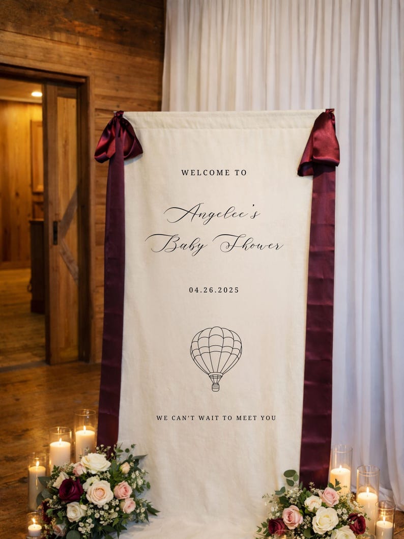 May include: A cream-colored baby shower welcome banner with burgundy ribbon accents. The banner reads "Welcome to Angelee's Baby Shower" with the date 04.26.2025 and a hot air balloon graphic. Floral arrangements and candles are at the base.