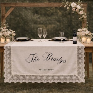 May include: A wooden table set for a wedding reception, with a white lace-trimmed table runner. The runner is embroidered with "The Bradys" and the date 03.20.2027. Champagne flutes, plates, and candles are arranged on the table.