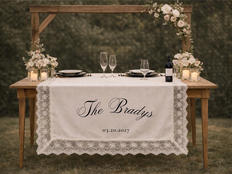 May include: A wooden table set for a wedding reception, with a white lace-trimmed table runner. The runner is embroidered with "The Bradys" and the date 03.20.2027. Champagne flutes, plates, and candles are arranged on the table.