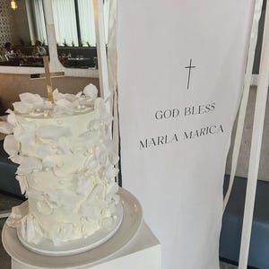 Pode incluir: Um bolo branco em camadas com decorações florais e detalhes de pérolas em um prato branco. Uma faixa com o texto "GOD BLESS MARLA MARICA" e uma cruz ao fundo. Uma cruz dourada no topo do bolo.